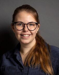 Porträt von Theresa Kunzelmann mit Brille und langen rötlich-blonden Haaren vor dunklem Hintergrund, sie lächelt in die Kamera.